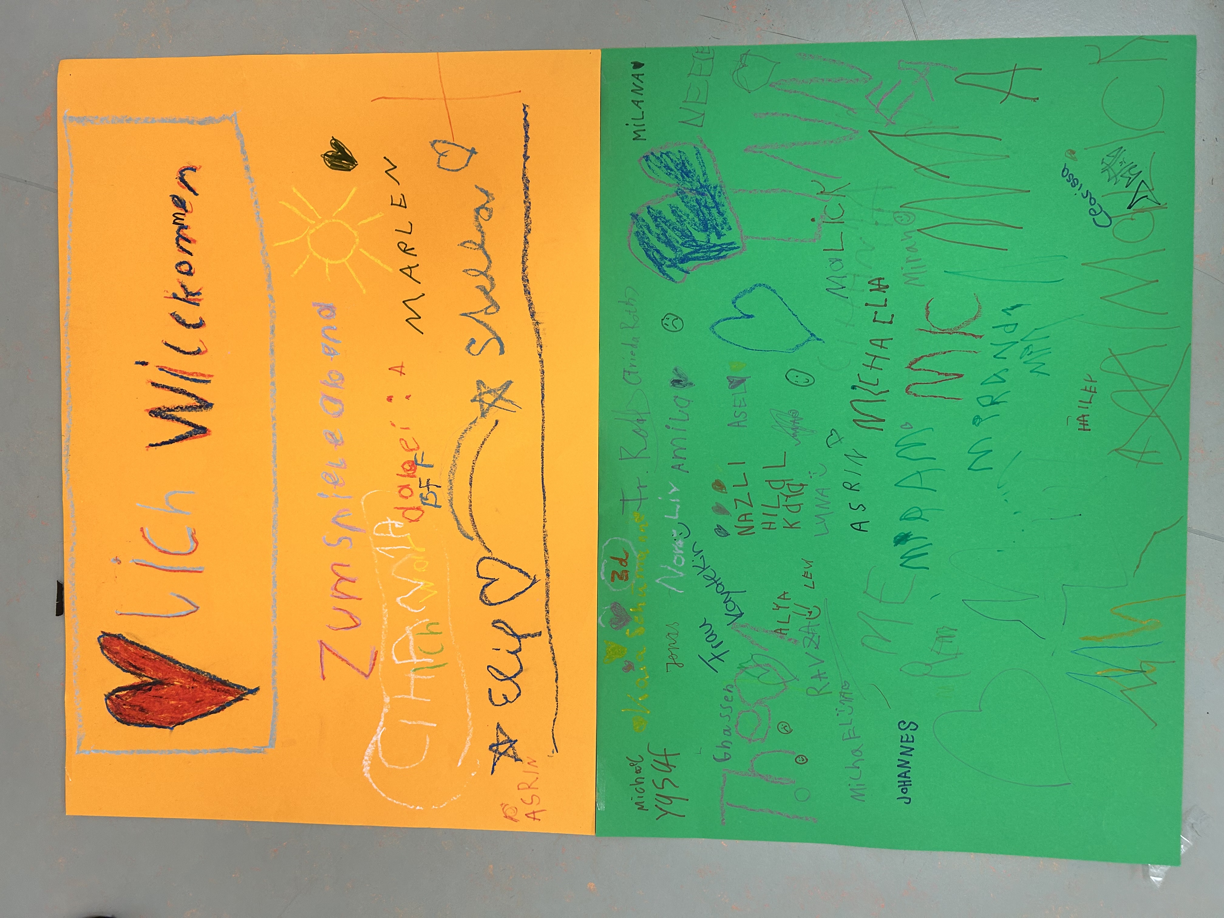 Selbst gestaltetes Willkommensplakat auf orangefarbenem und grünem Papier. Darauf steht „Herzlich Willkommen“ sowie „Zum Spieleabend“; zahlreiche Kinder haben ihre Namen, Herzen, Sterne und weitere kleine Zeichnungen darauf hinterlassen.