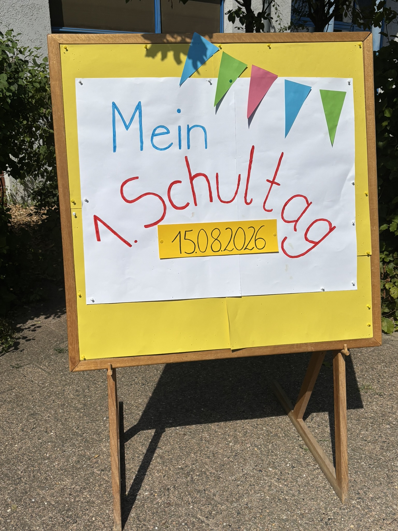Auf dem Foto ist eine bunt geschmückte Schultafel vor dem Schulgebäude zu sehen. Auf einem großen weißen Plakat steht in bunten Buchstaben „Mein Schul…“, darunter das Datum 15.08.2026. Bunte Wimpel schmücken den oberen Rand. Die sommerliche Gestaltung vermittelt eine fröhliche und festliche Stimmung passend zur Einschulung.